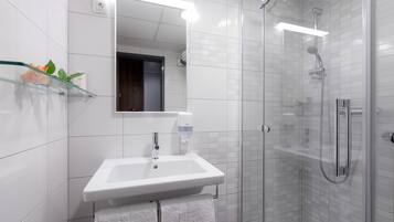 A modern bathroom with a glass shower enclosure, a white sink with a soap dispenser, and a glass shelf with a plant.