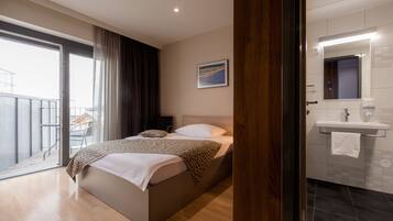 A modern hotel room with a large bed, a balcony, a bathroom with a mirror and sink, and a view of the city.