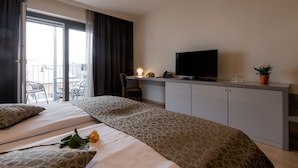 A hotel room with a bed, a desk with a TV, a potted plant, and a balcony with chairs.