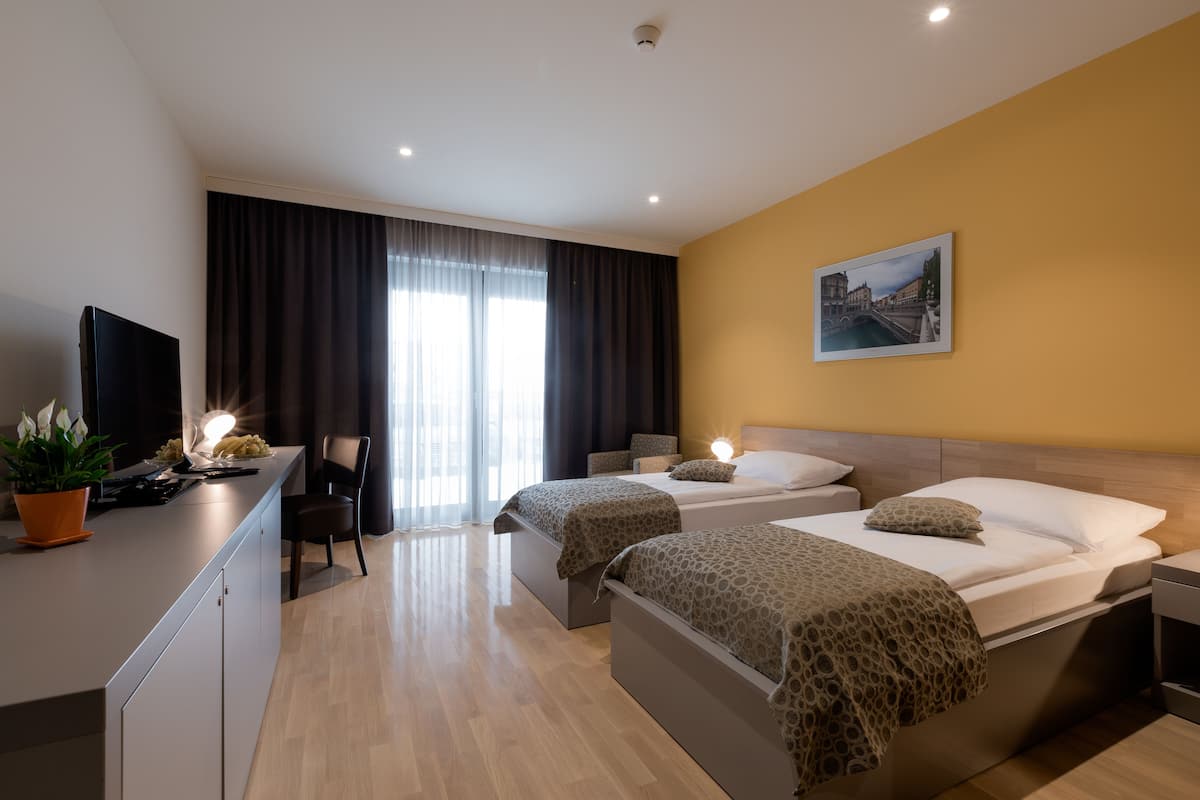 Habitació d'hotel moderna amb dos llits, una taula de treball amb televisió de pantalla plana, una planta en test i una finestra gran amb cortines.