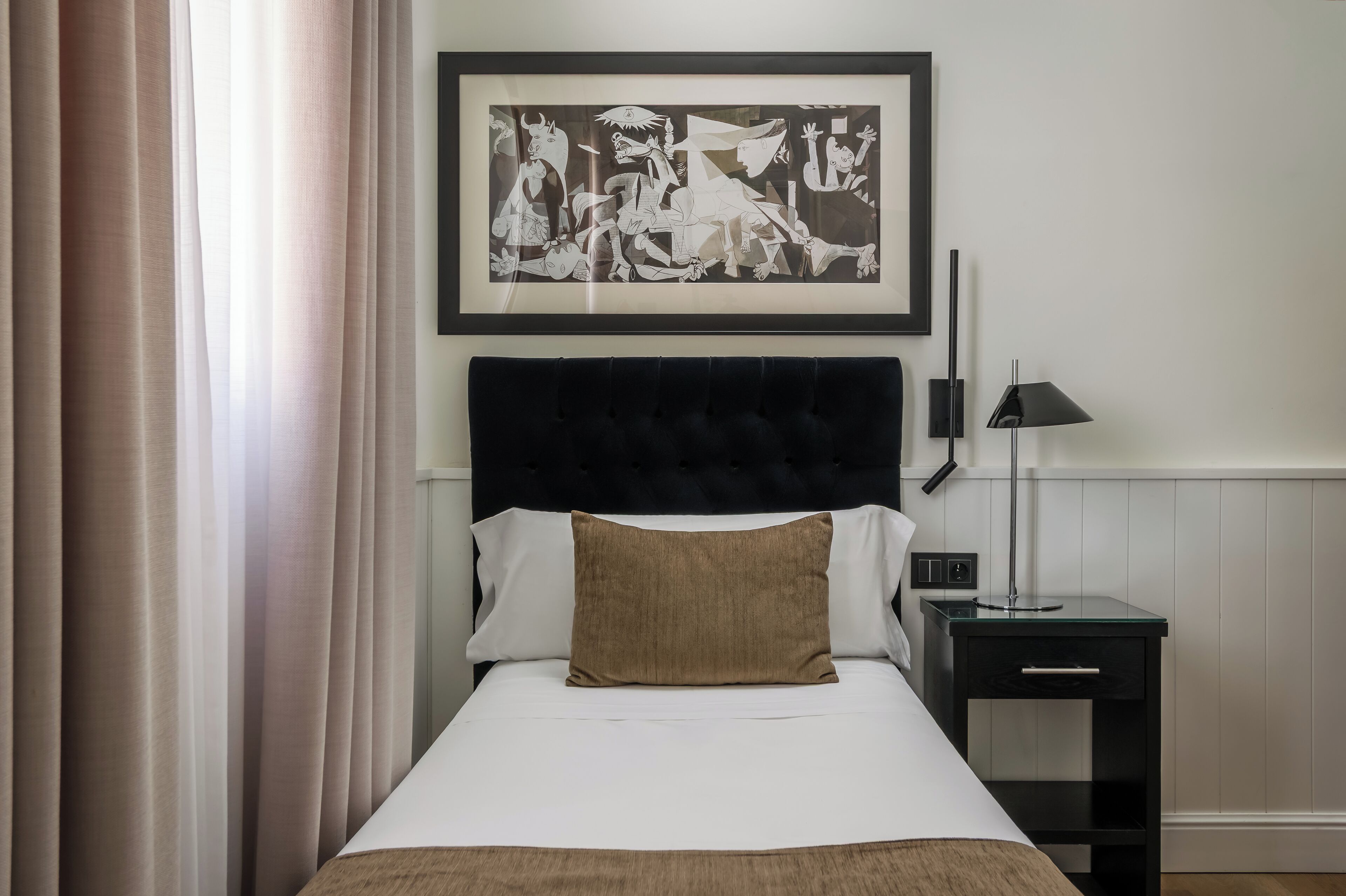 A neatly made bed with a black headboard, a framed artwork above it, and a bedside table with a lamp.