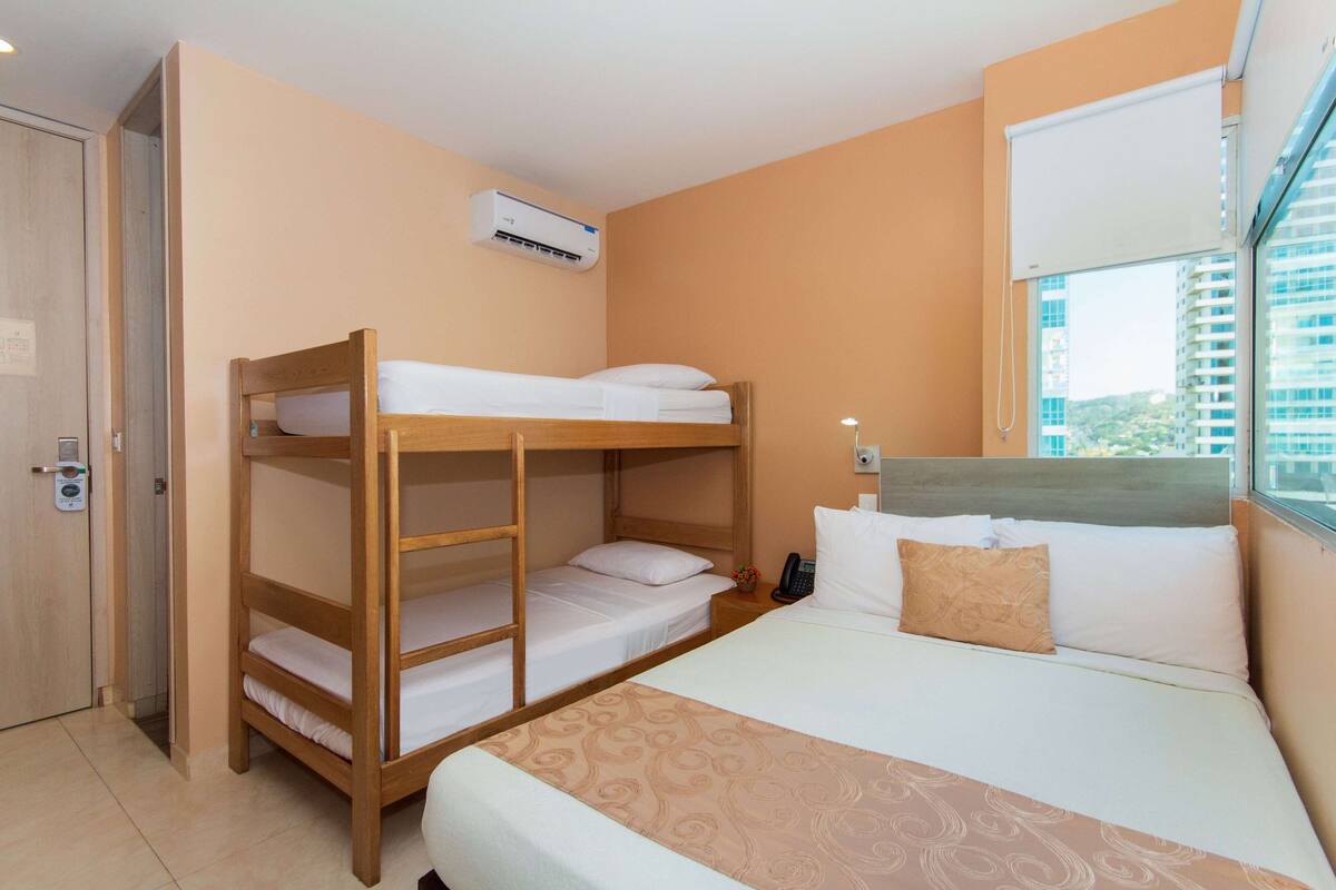 Habitación de hotel con una litera, una cama individual, aire acondicionado, una mesita de noche con teléfono y una ventana con vista a edificios.