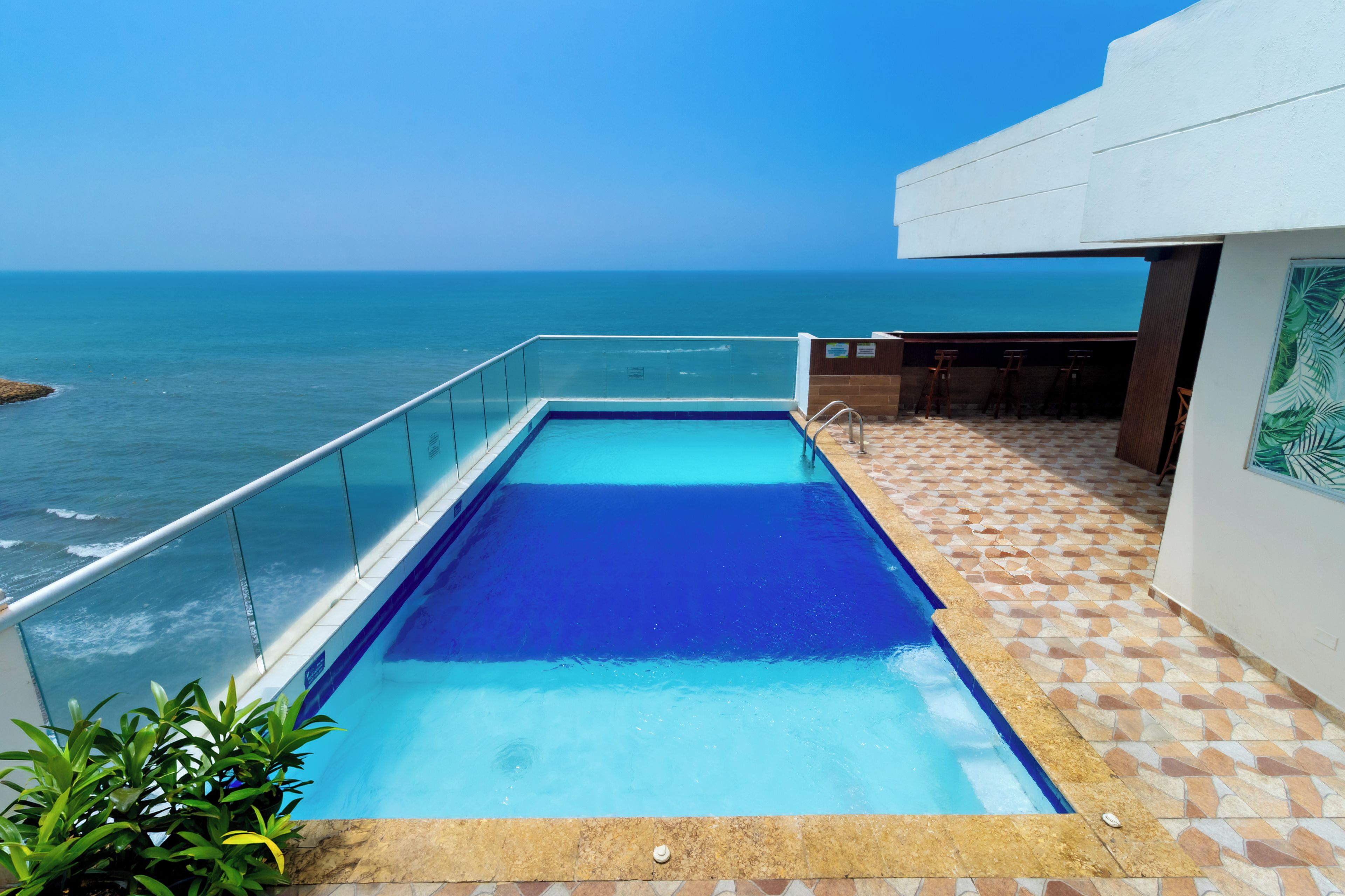 Una piscina rectangular con superficie de agua azul cristalina, rodeada por una barandilla de vidrio y un área de terraza con baldosas y vistas al océano.