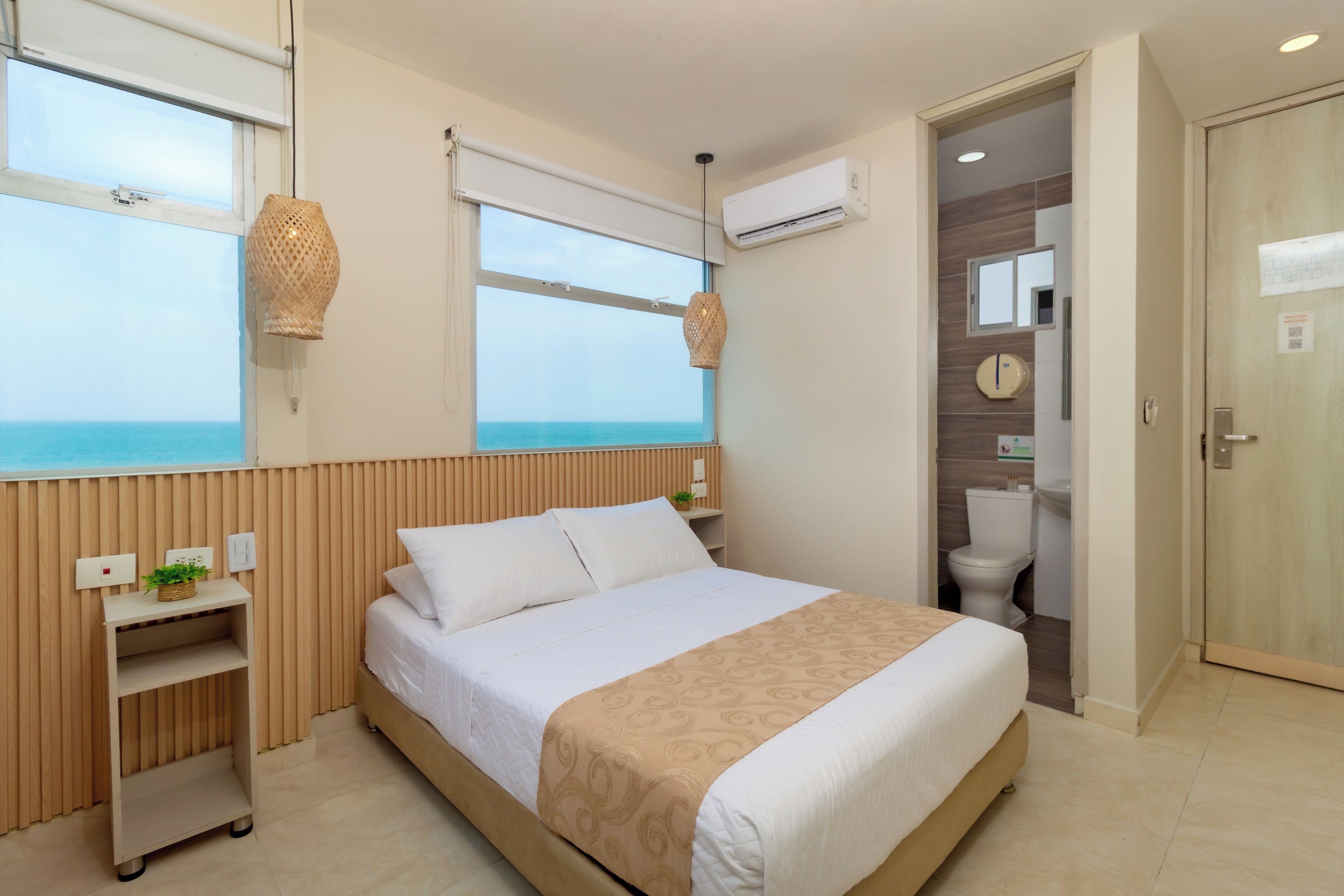 A hotel room with a bed, a window with a view of the sea, an air conditioning unit, and a bathroom with a toilet and sink.