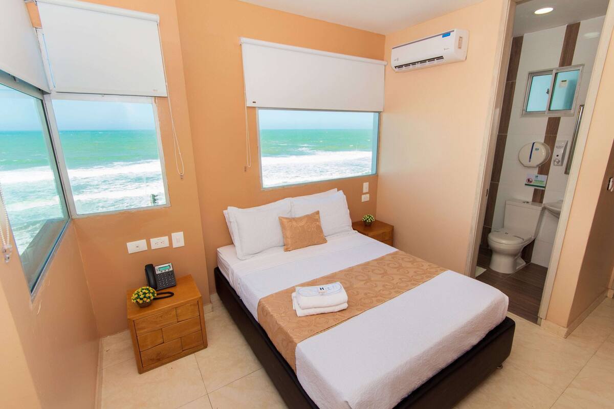 Un dormitorio con cama, una mesita de noche, una ventana con vistas al océano y un baño visible a través de una puerta abierta.
