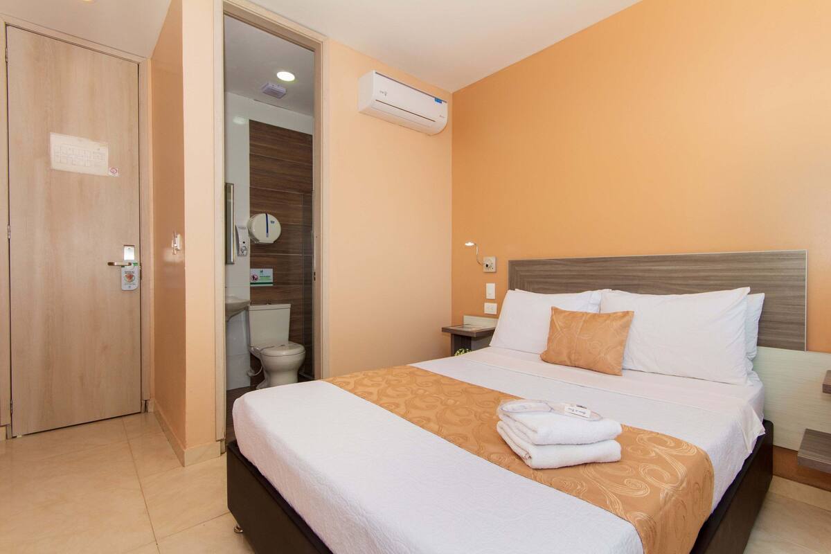 Una habitación de hotel con cama, mesita de noche, equipo de aire acondicionado, una puerta y un baño visible a través de una puerta abierta.