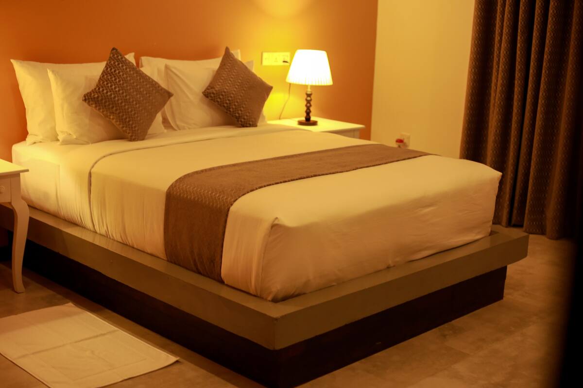 A neatly made bed with white linens and a brown throw, a bedside lamp, and a nightstand with a lamp.