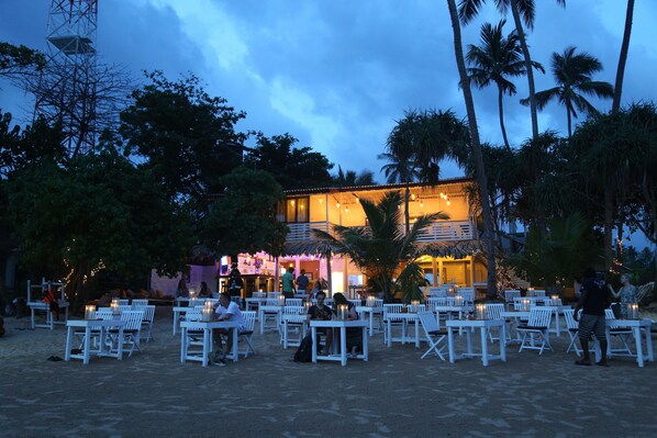 An outdoor seating area with white tables and chairs, palm trees, and a building with warm lighting in the background. - Citrine Mirissa (Mirissa)