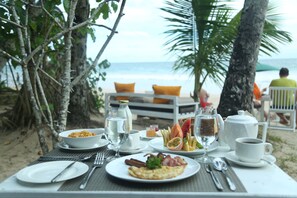 A breakfast table with plates of food, a cup of coffee, and a bottle of water on a beach with palm trees and a view of the ocean. - Citrine Mirissa (Mirissa)