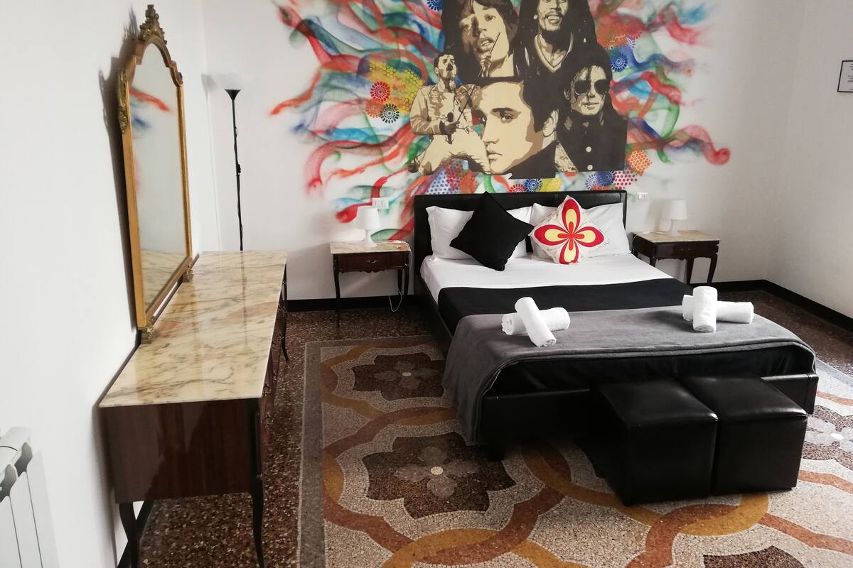 A hotel room with a bed, a nightstand, a mirror, a wall art, and a patterned rug.