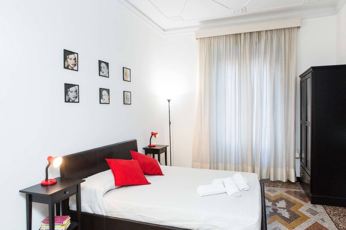 A neatly arranged hotel room with a bed, a nightstand with a lamp, a wardrobe, and a wall with framed pictures.