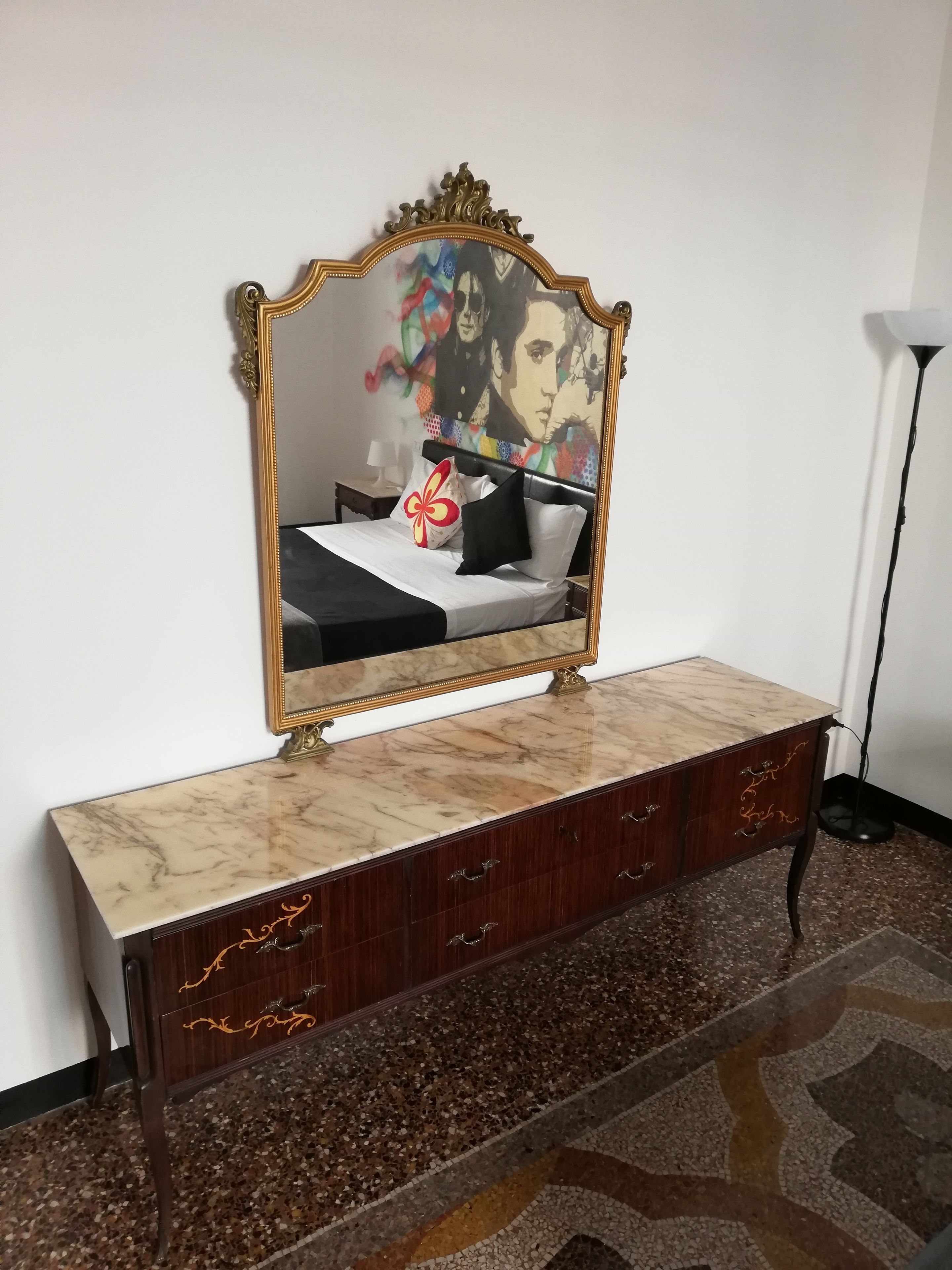A bedroom with a bed, a marble bedside table, a dark wooden dresser with drawers, a gold-framed mirror, and a floor lamp.