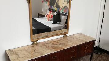 A bedroom with a bed, a marble bedside table, a dark wooden dresser with drawers, a gold-framed mirror, and a floor lamp.