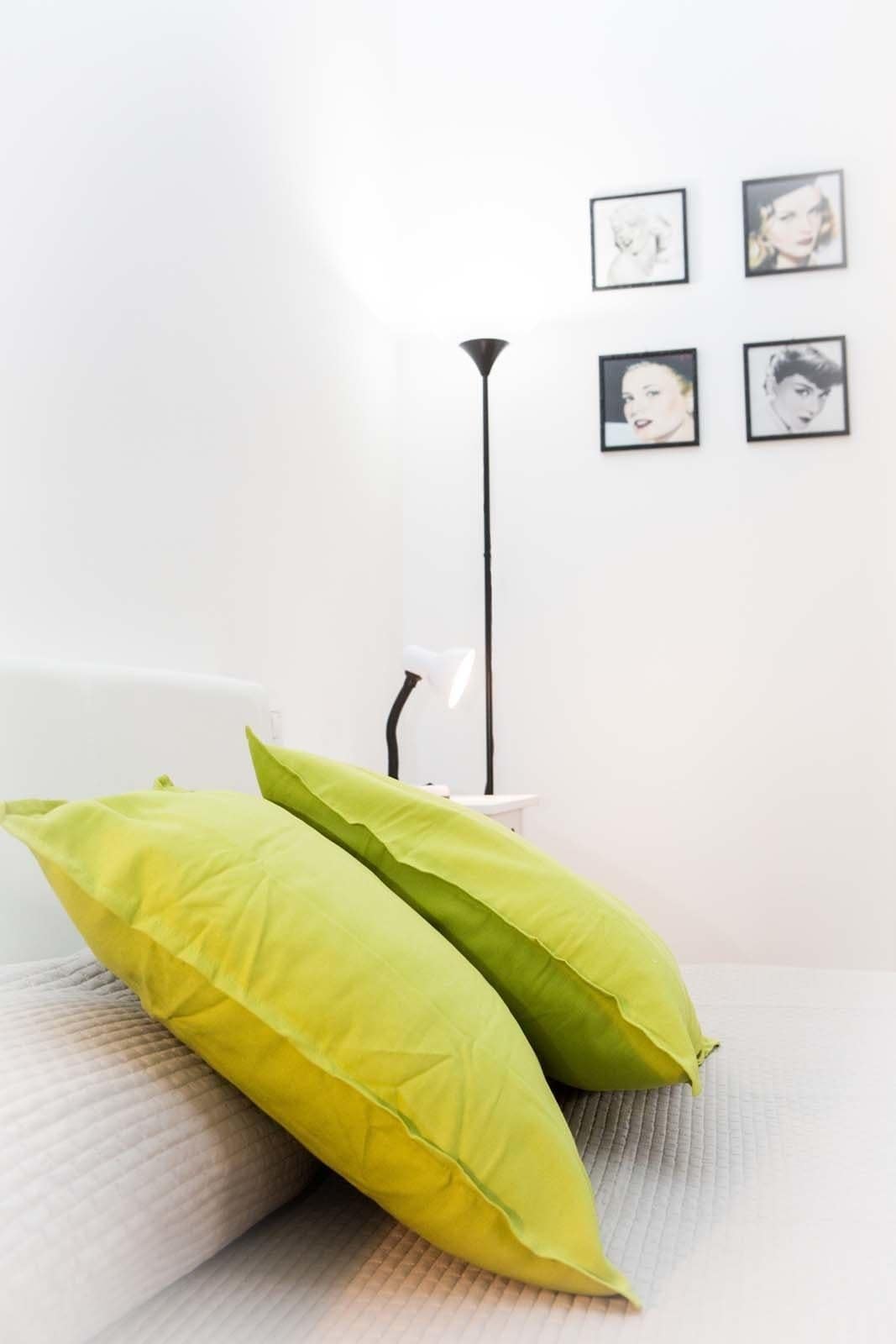 A room with a white wall, a floor lamp, and four framed pictures. There are two green pillows on a white couch.