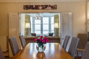 A dining room with a wooden table, chairs, a vase of flowers, and a view through a bay window.