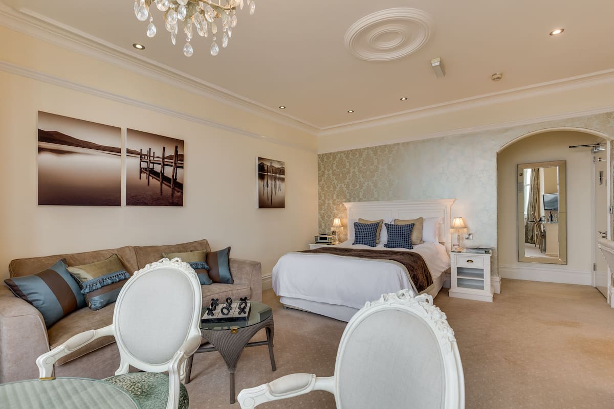 A hotel room with a bed, a sitting area with a chair and a small table, a mirror, and artwork on the wall.