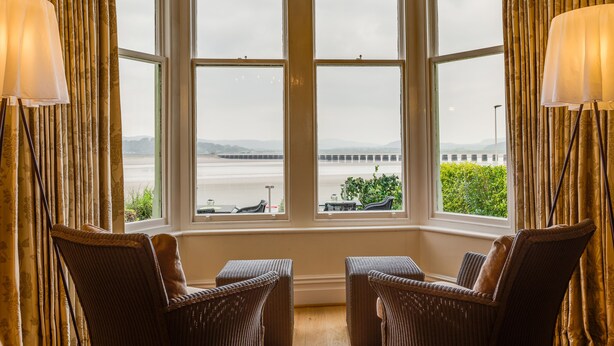 A room with a bay window overlooking a body of water and a bridge, featuring wicker chairs and a lamp. - Number 43 (Carnforth)