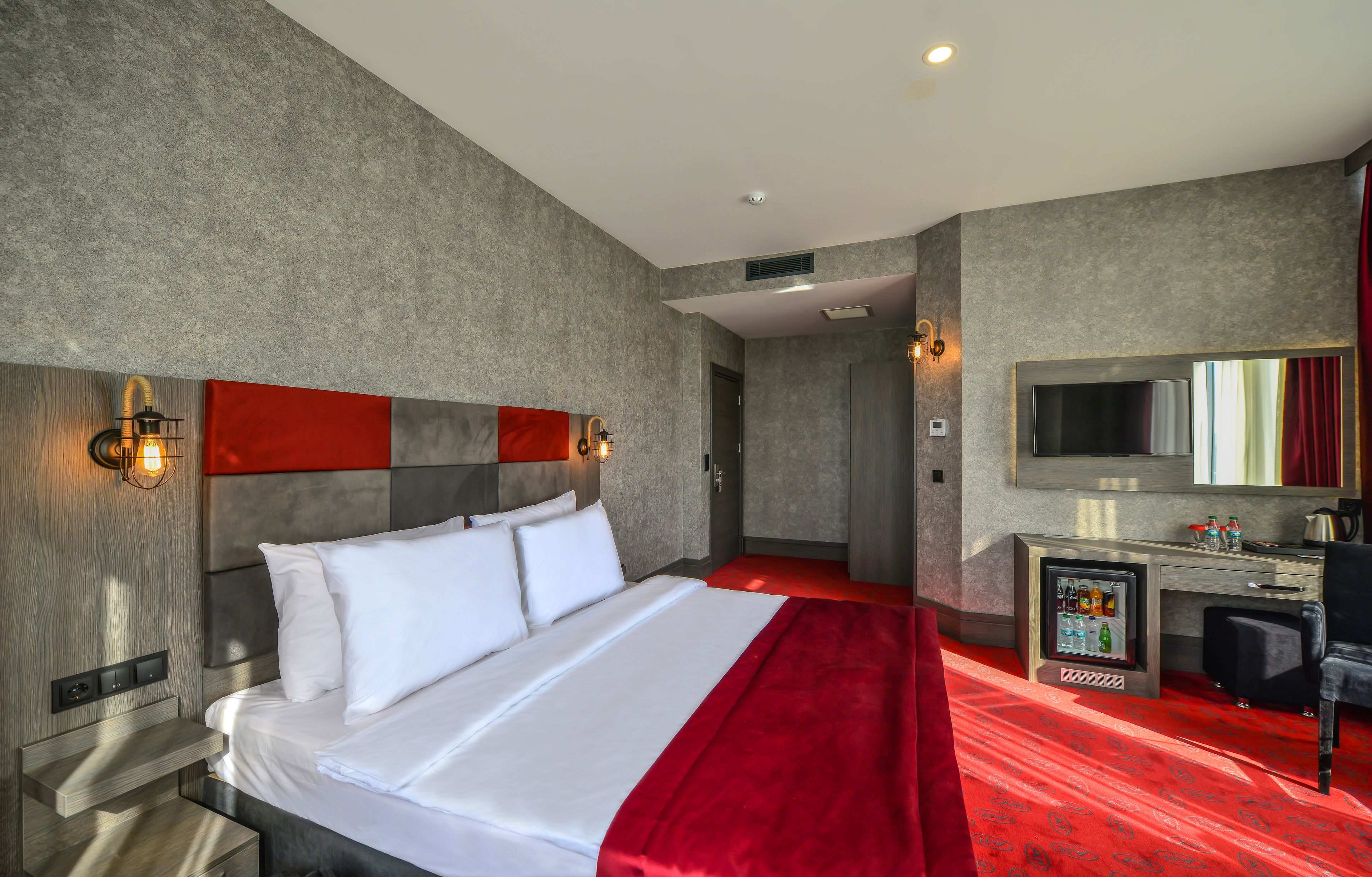 A modern hotel room with a large bed, a desk with a TV, and a minibar.