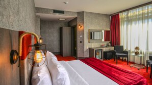 A modern hotel room with a large bed, a desk, and a chair. There is a mirror on the wall and a window with curtains.
