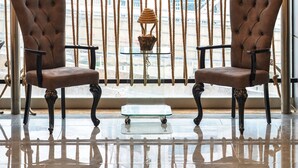 A room with two brown tufted chairs, a glass coffee table, and a view of a cityscape through a window with a rope curtain.