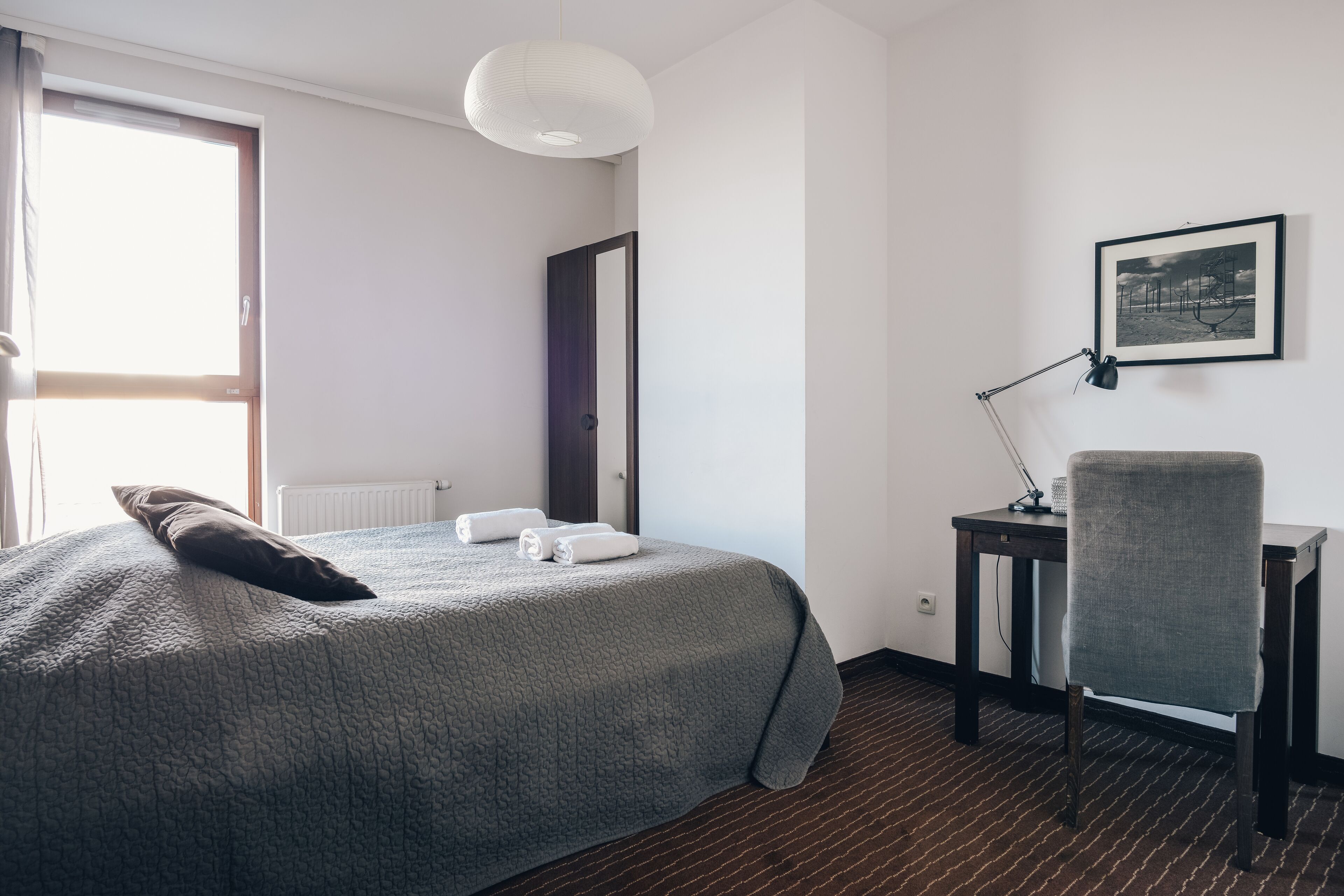Quarto de hotel com cama, escrivaninha, cadeira, abajur e um quadro na parede.