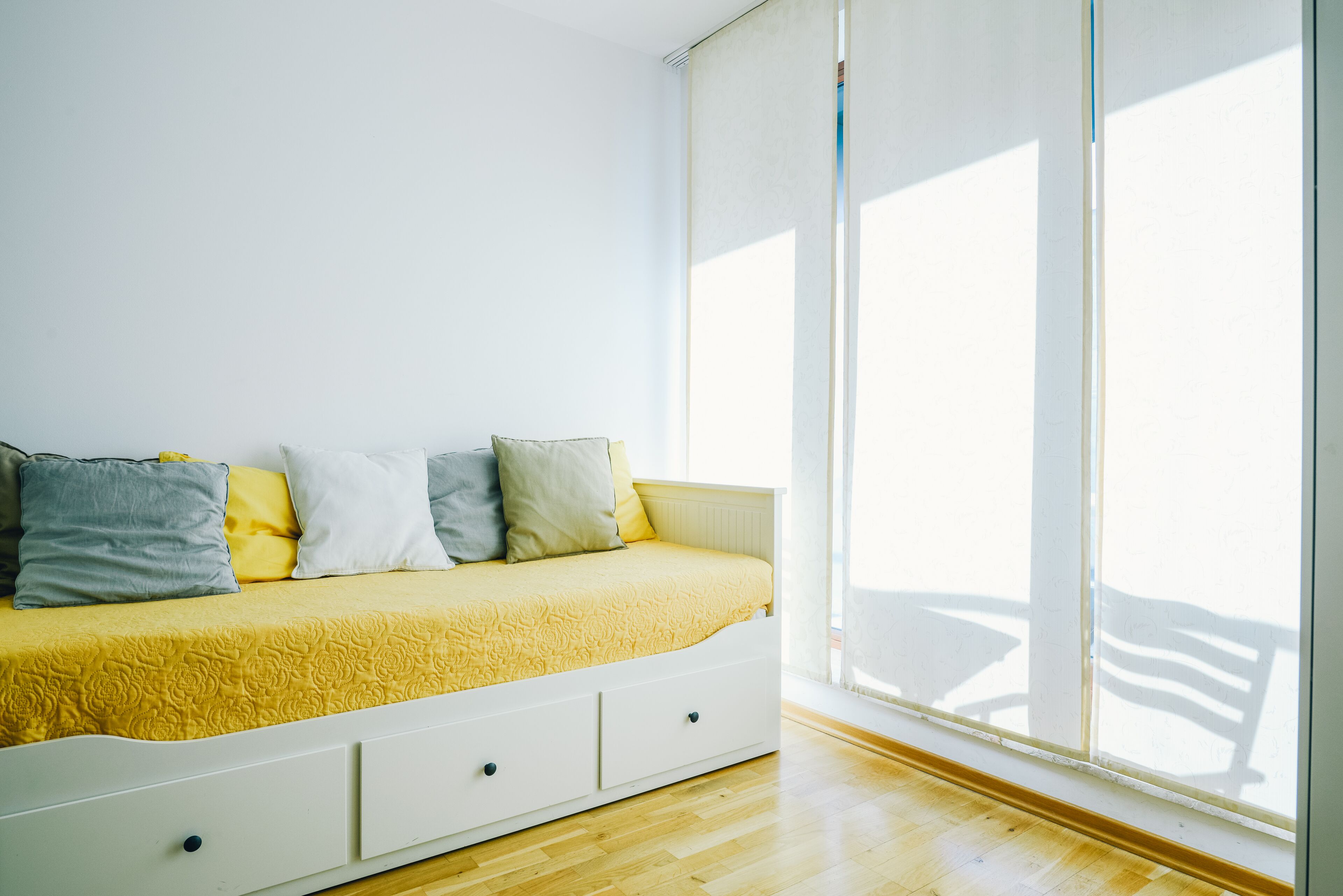 A bright room with a bed against a white wall, a large window, and wooden flooring.