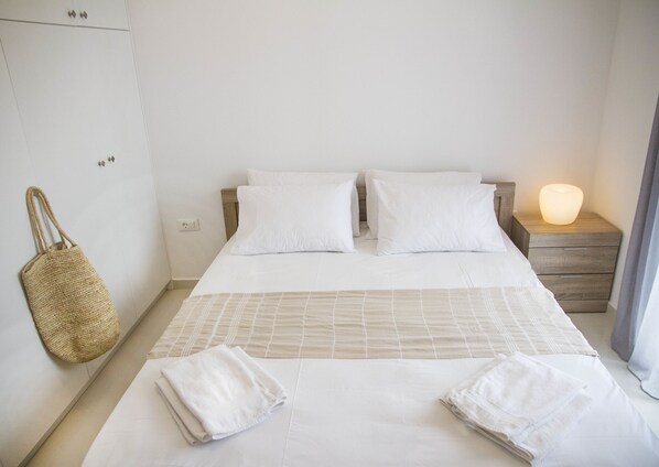 Una cama bien hecha con sábanas blancas, un bolso tejido colgado en la pared y una mesita de noche con una lámpara.