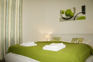 Un lit bien fait, avec une couverture verte et des oreillers blancs, un rideau à rayures et une œuvre d’art accrochée au mur.