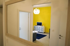 Une pièce avec une porte blanche, un téléviseur posé sur un meuble blanc, un tapis bleu et un mur jaune.