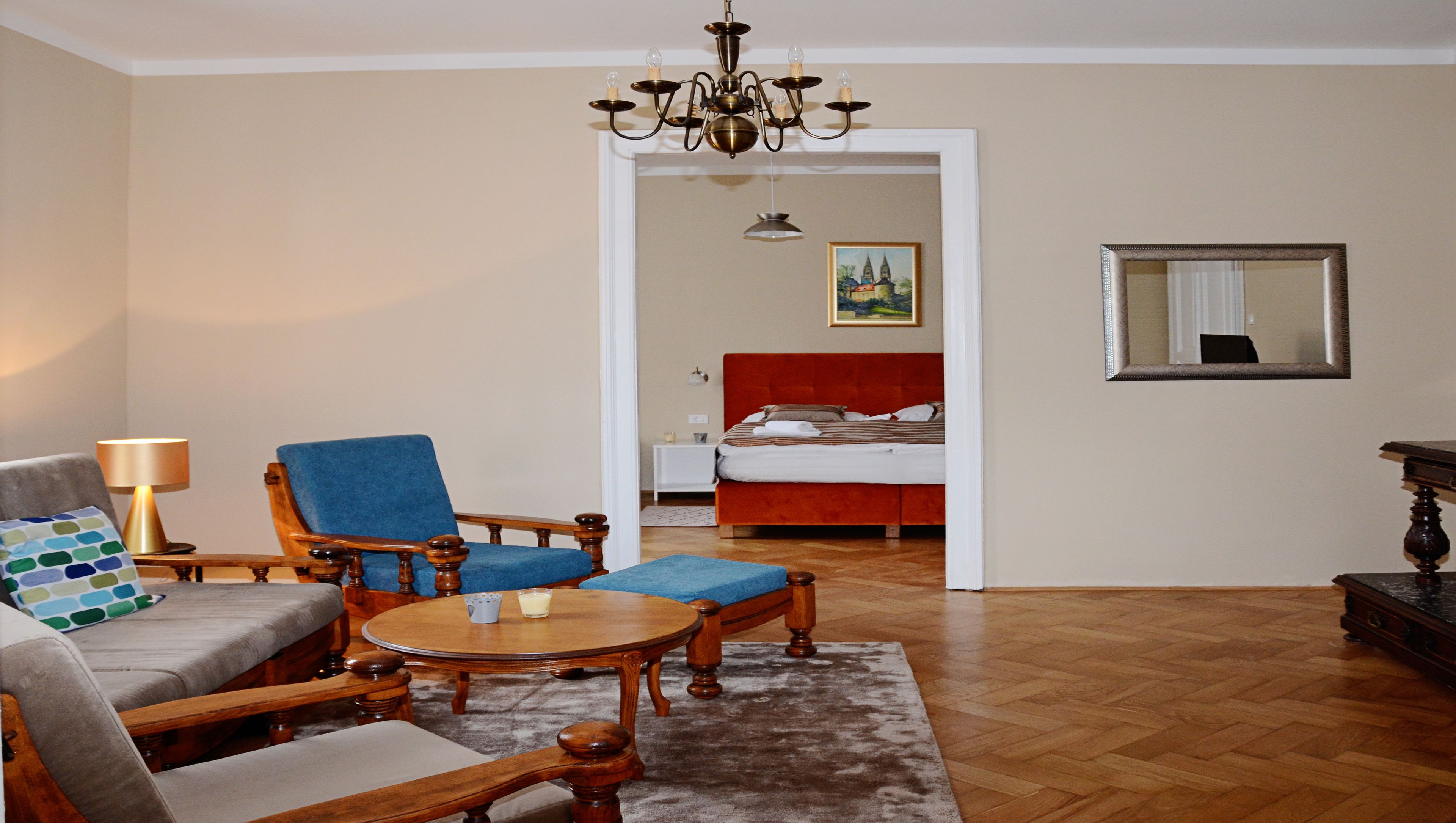 Een woonkamer met een bank, fauteuil en salontafel. Achterin staat een bed met een rode hoofdeinde.