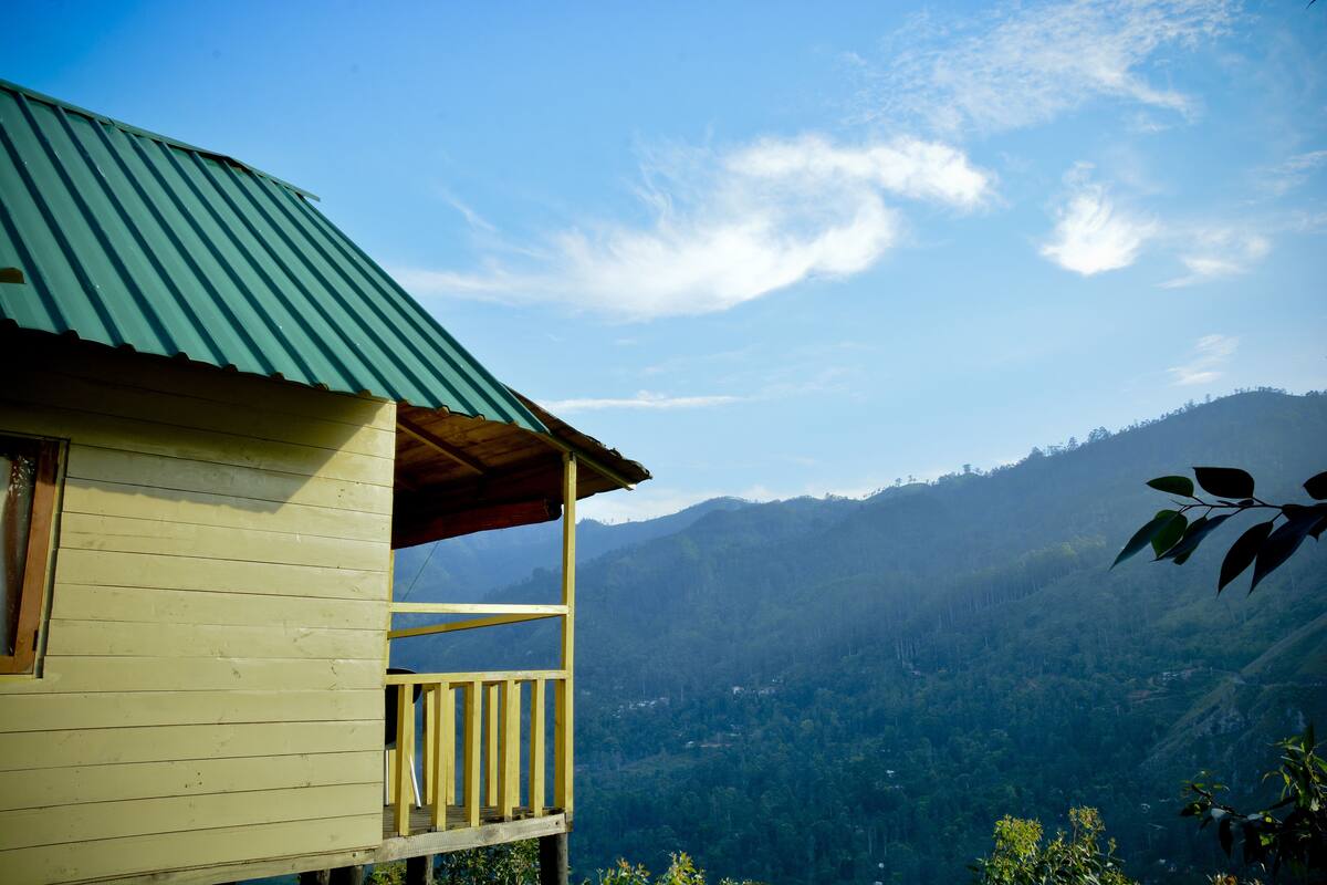 A wooden cabin with a green roof, a balcony, and a view of a forested mountain range.