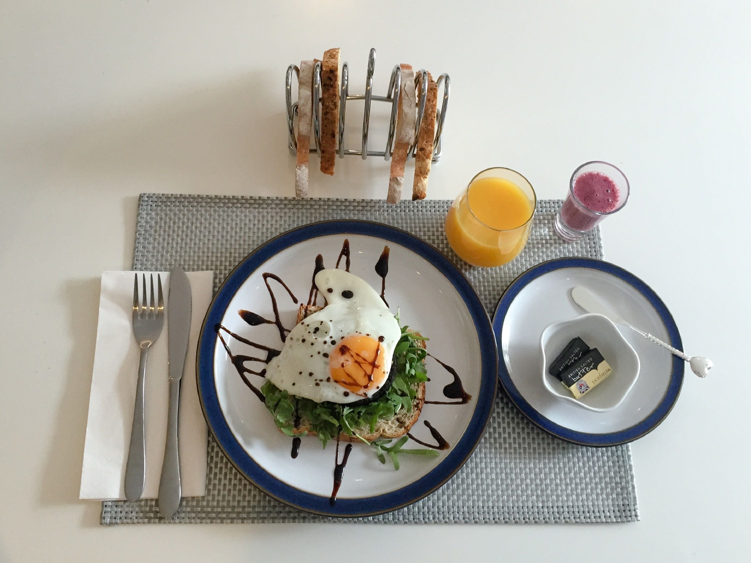 Un plato de desayuno con un huevo frito, verduras, tostadas, un vaso de jugo de naranja y un batido, sobre una mesa con posavasos.