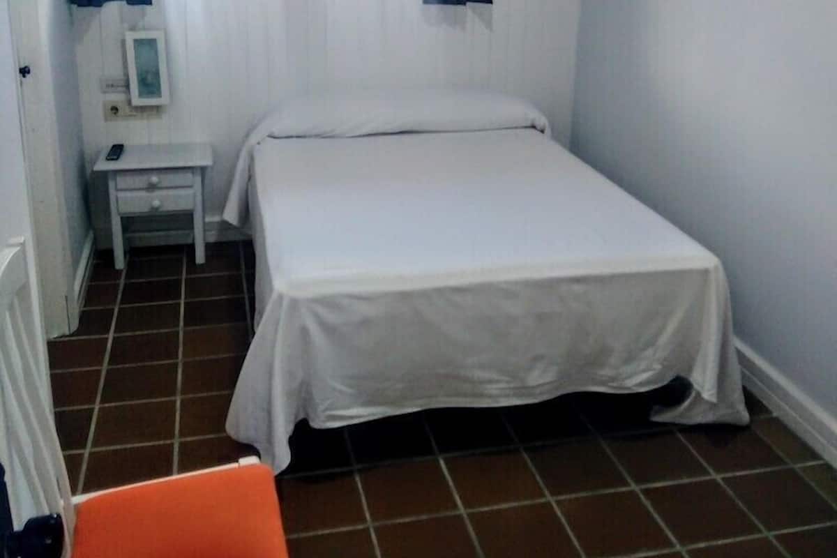 Een kleine, schone kamer met een bed, nachtkastje, stoel en een raam met gordijnen.