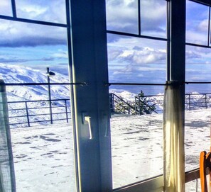 Vista a través de una ventana con barandilla de metal, suelo cubierto de nieve y una montaña al fondo.