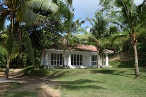 Un bâtiment blanc avec un toit de chaume, entouré de palmiers et d’une zone herbeuse.