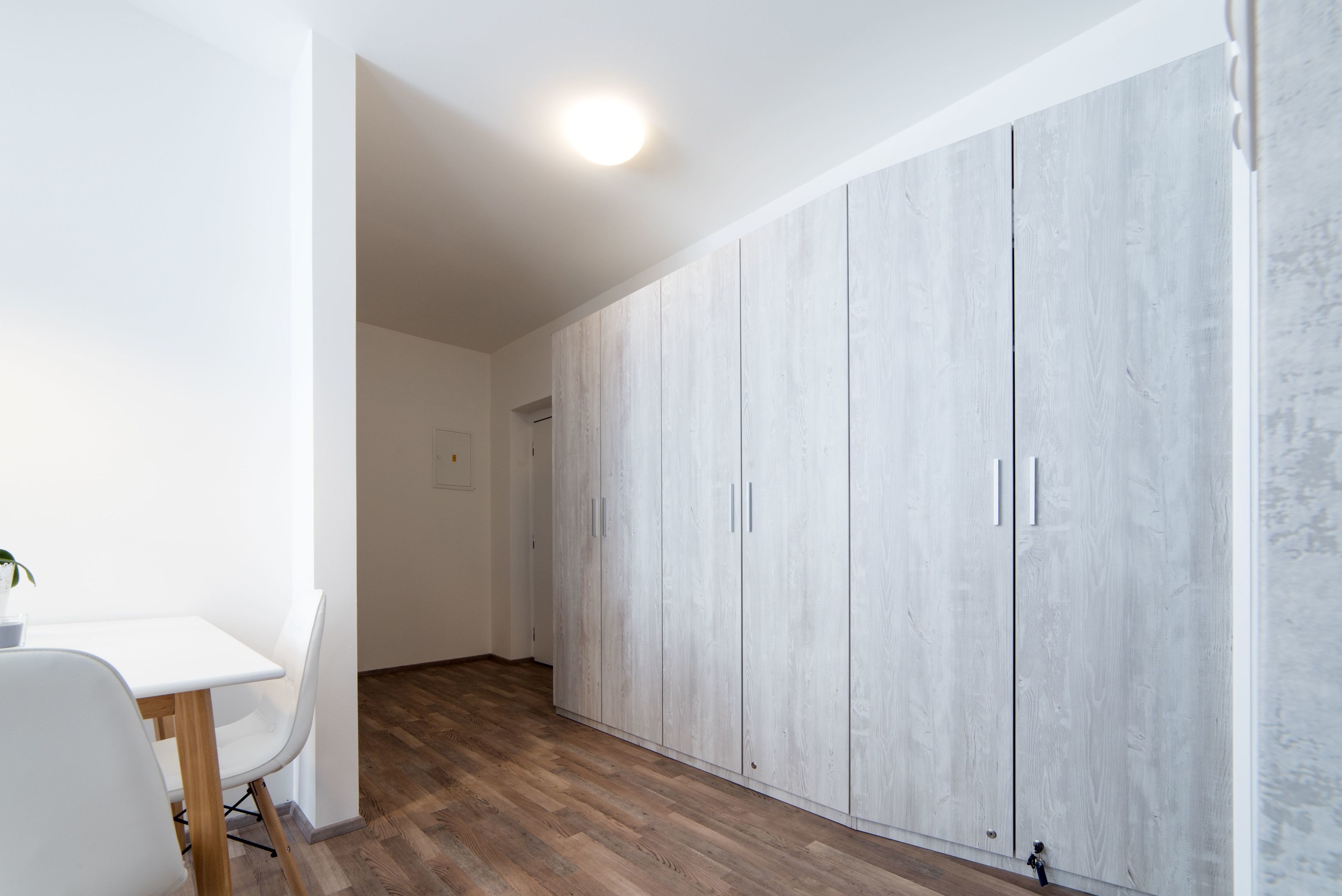 A room with a white desk, white chairs, a wooden floor, and a large wardrobe with multiple doors.
