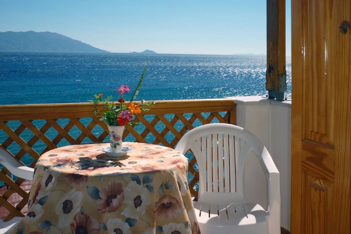 A table with a floral tablecloth, a vase with flowers, and a chair on a balcony overlooking the sea.