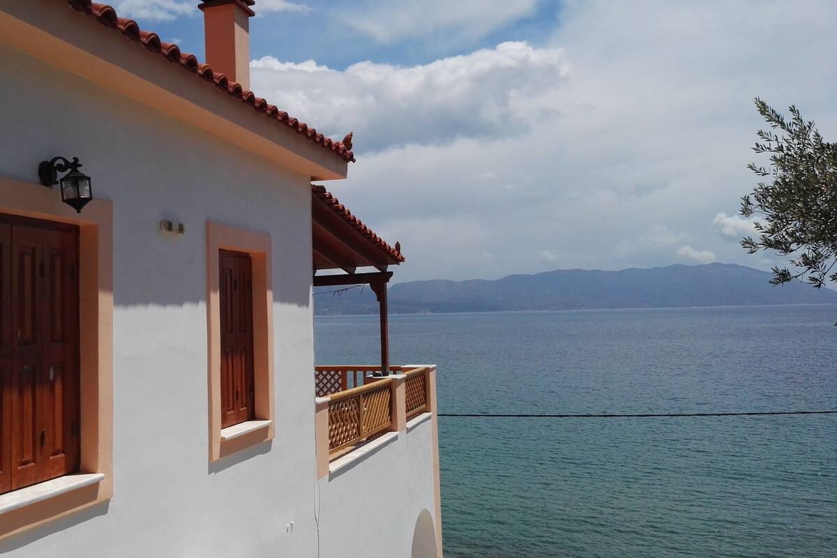 A white building with wooden doors and windows, a balcony with a wooden railing, and a view of a calm sea and distant mountains.