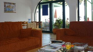 Ein Zimmer mit zwei orangefarbenen Sofas, einem Couchtisch mit Zeitschriften und einer Blumenvase, einer Weltkarte an der Wand und Blick ins Freie durch eine Glastür.