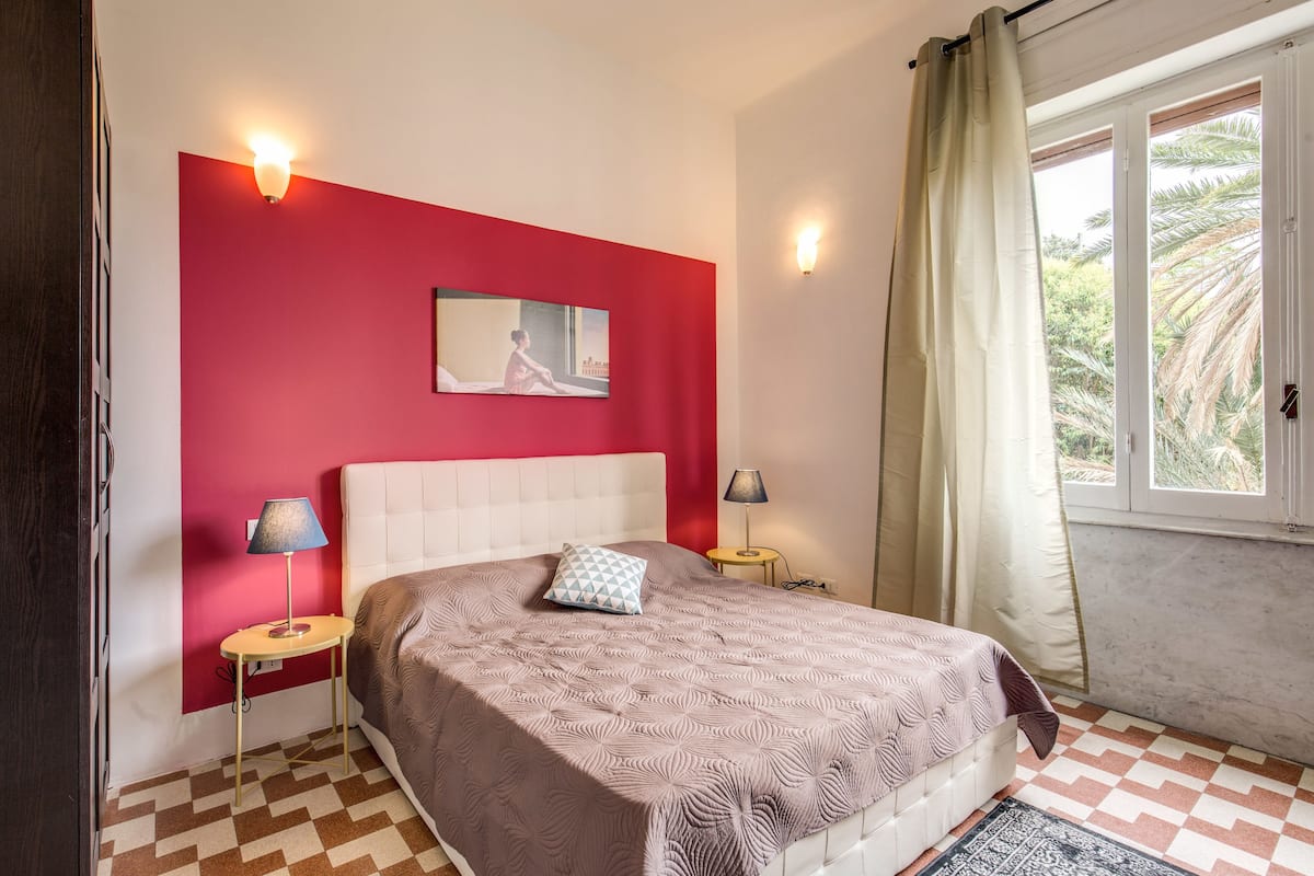 A bedroom with a checkered floor, a white bed, a red accent wall, and two bedside lamps.