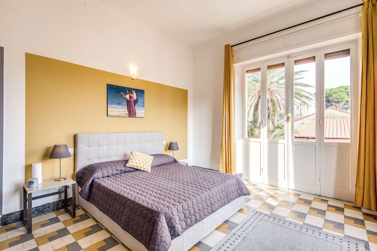 A bedroom with a bed, a nightstand, a lamp, a painting, and a view through a sliding door.