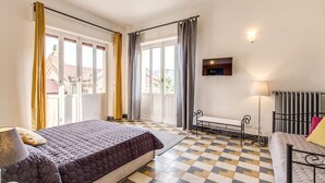 Una habitación de hotel con piso a cuadros, una cama con cubrecama morado, un banco, un radiador y un televisor fijado a la pared.