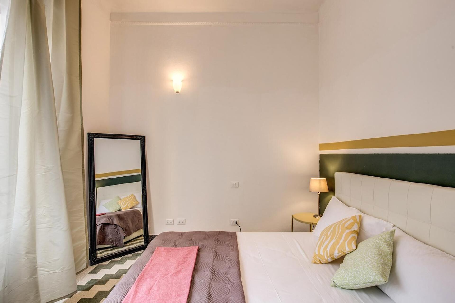 Een hotelkamer met een bed, nachtkastje, lamp, spiegel en een raam met gordijnen.