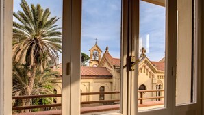 Vista a través de una ventana con barandilla de balcón, palmeras y un edificio con una torre de reloj.