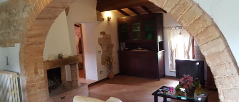 A cozy living area with a stone archway, a fireplace, a sofa, a glass coffee table, and a kitchen area visible through the archway.