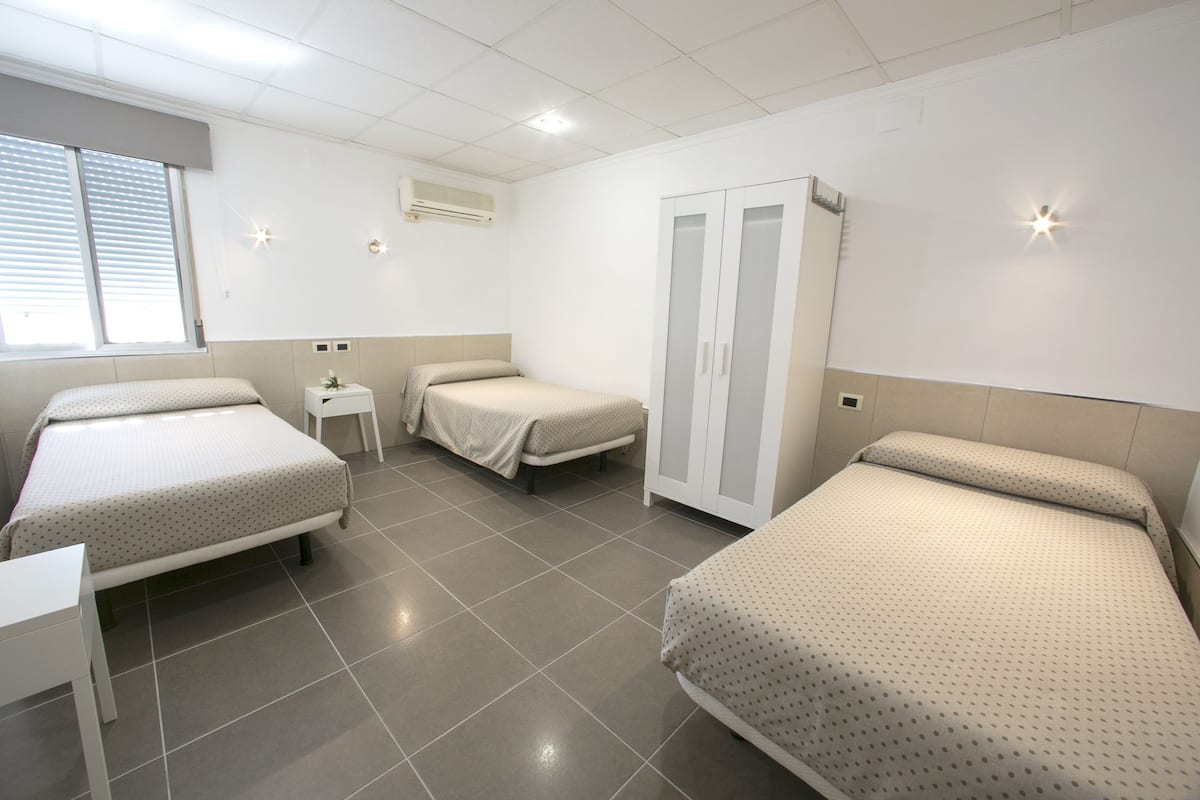 Ein Zimmer mit drei Betten, einem Kleiderschrank, einem kleinen Tisch und einem Fenster mit Jalousien.