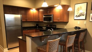 A kitchen with wooden cabinets, a stainless steel refrigerator, a microwave, and a stove. There are two bar stools with patterned cushions. - Siebkens Resort (Elkhart Lake)