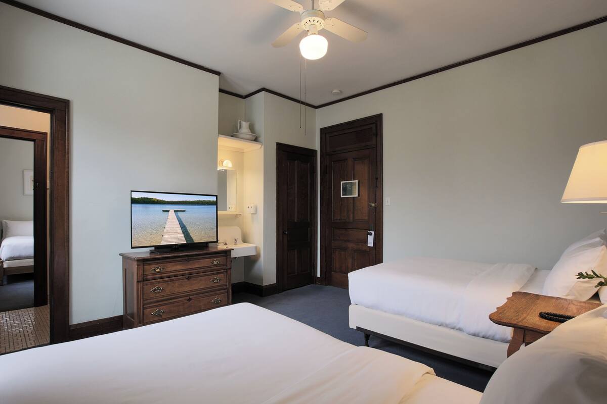 A hotel room with a bed, a dresser, a television displaying a scenic view, a wooden door, and a nightstand with a lamp.
