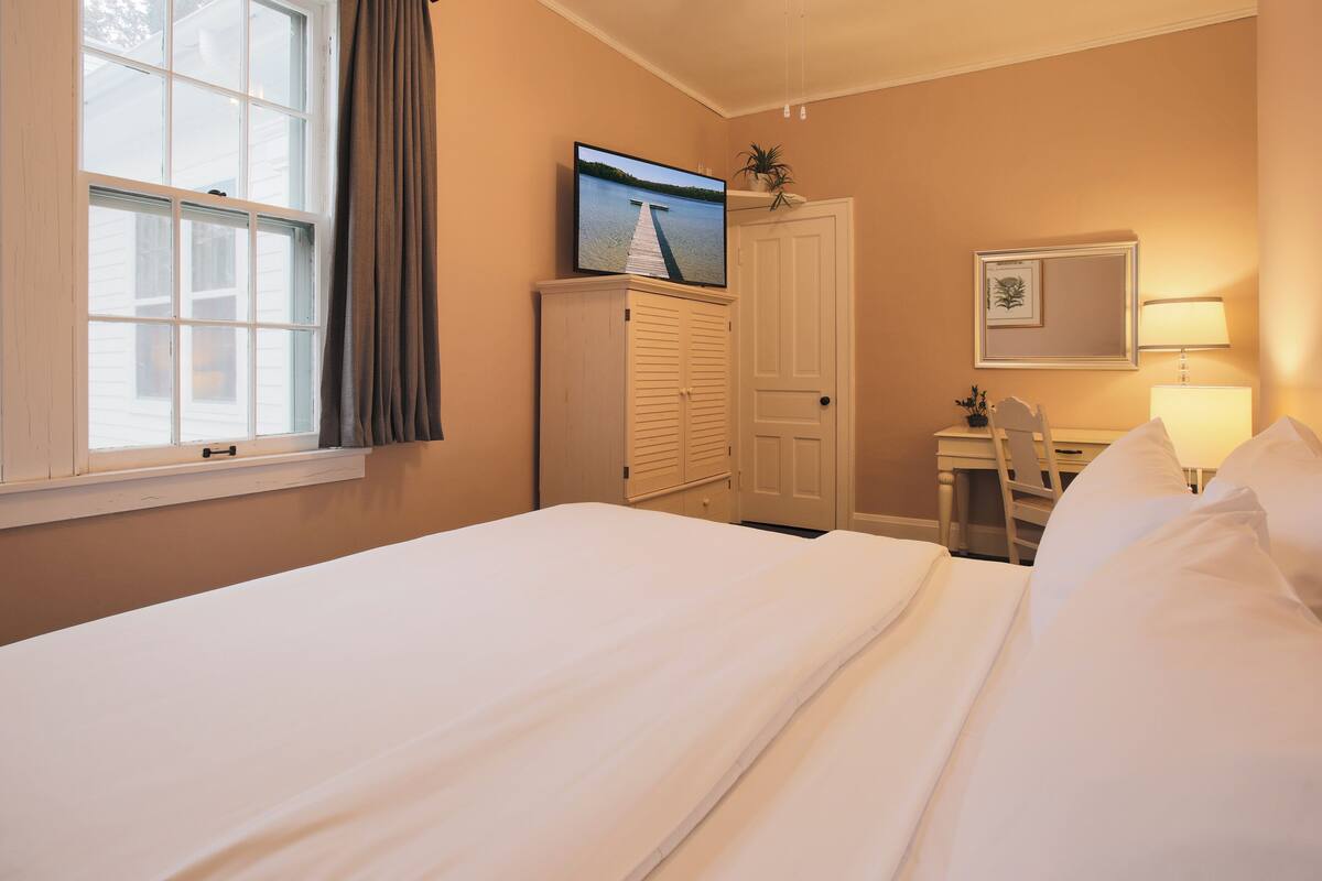 A hotel room with a large bed, a desk with a chair, a wardrobe, a television on a stand, and a window with curtains.
