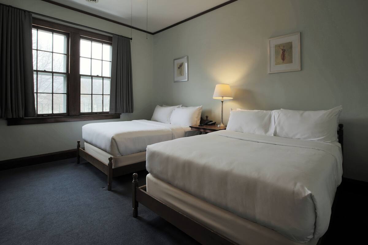 A hotel room with two beds, a window with curtains, a bedside lamp, and two framed pictures on the wall.